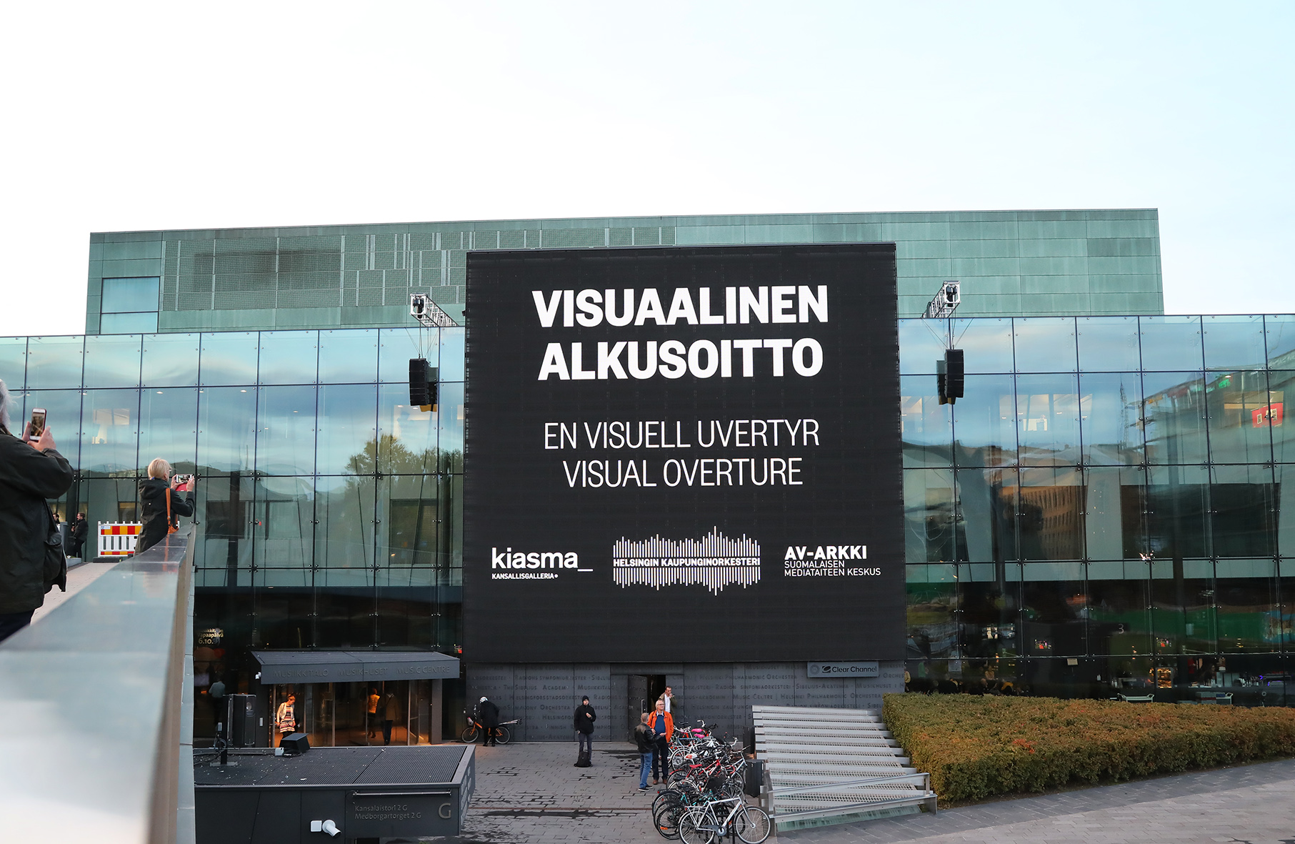 Kuvassa näkyy Helsingin Musiikkitalon seinä ja seinällä oleva ulkonäyttö. Näytöllä näkyy Visuaalinen alkusoitto -mediataideprojektin nimi ja sen järjestäjät Kiasma, Helsingin kaupunginorkesteri ja AV-arkki. Kuvan on ottanut Maarit Kytöharju.