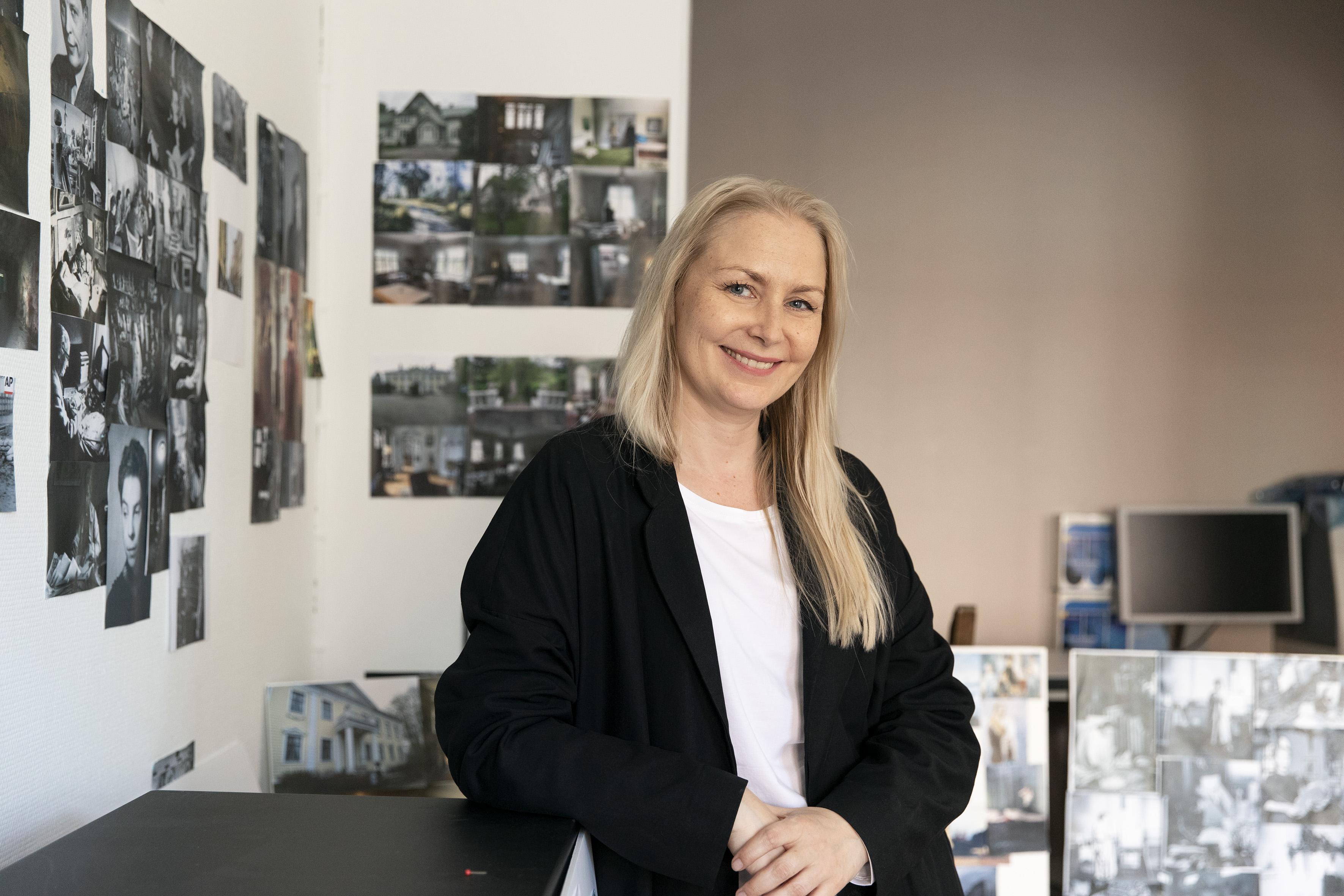 Kuvassa Zaida Bergroth nojaa tasoon ja katsoo suoraan kameraan hymyillen. Taustalla näkyy seinään kiinnitettyjä erilaisia valokuvia.