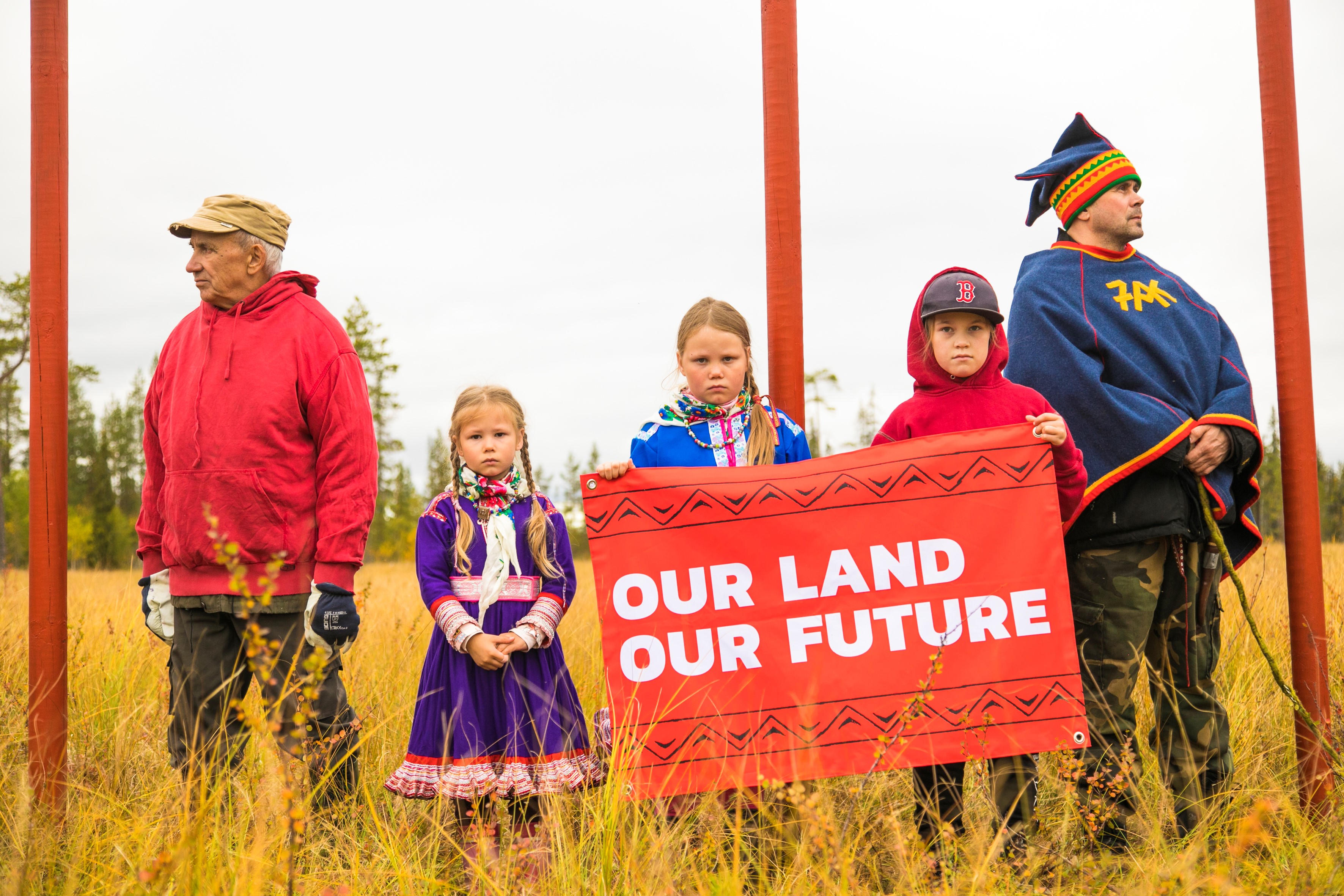 Kuvassa reunoilla kaksi aikuista ja keskellä kolme lasta peltomaisemassa. Lapset katsovat suoraan kameraan ja pitelevät kylttiä, jossa lukee "Our land, our future".