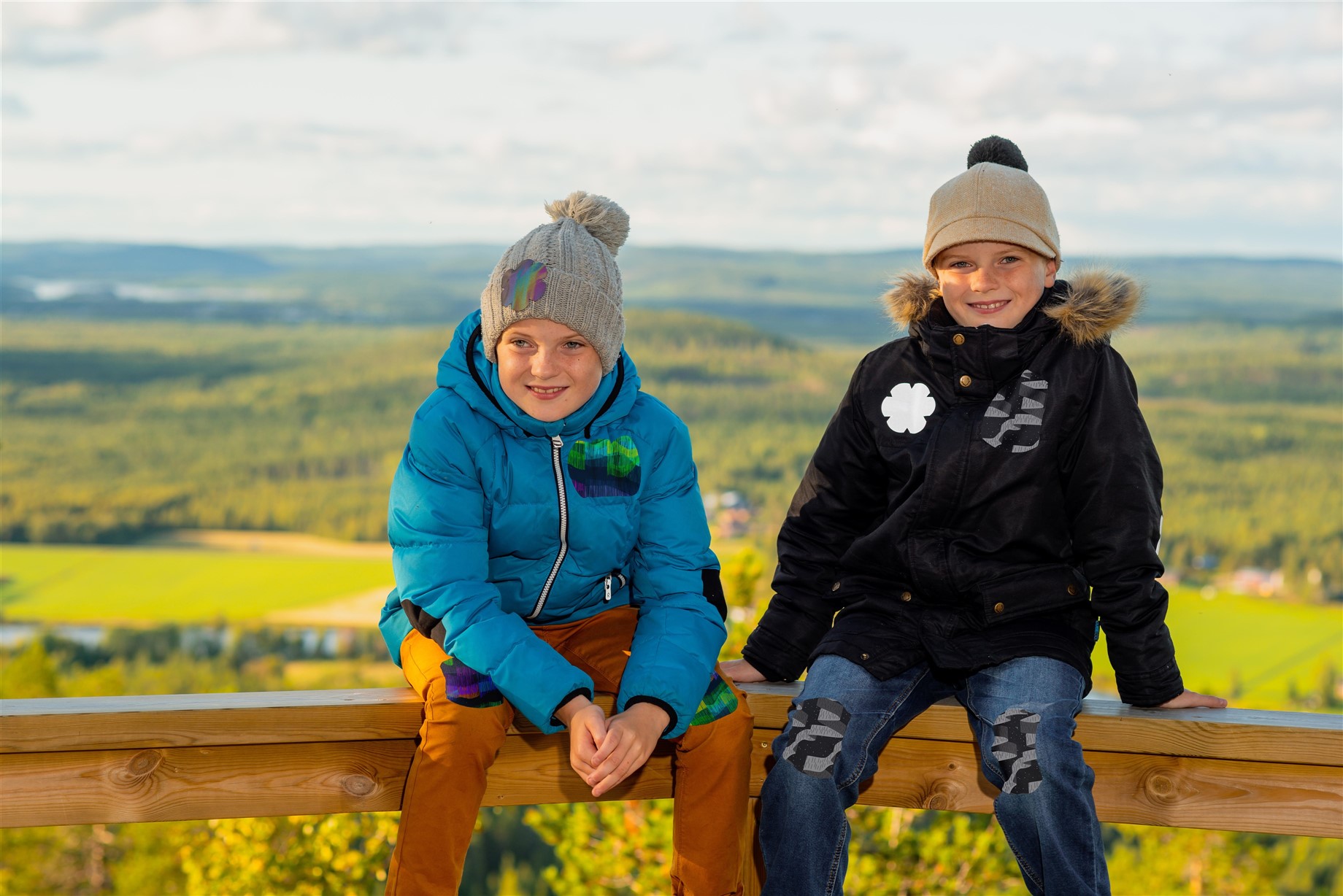 Kuvassa kaksi poikaa istuu kaiteella syksyisessä peltomaisemassa. Heillä on yllään syys- tai talvivaatteet ja vaatteissa on Oikiat Designin kehittämiä heijastavia Vaatelaastareita.