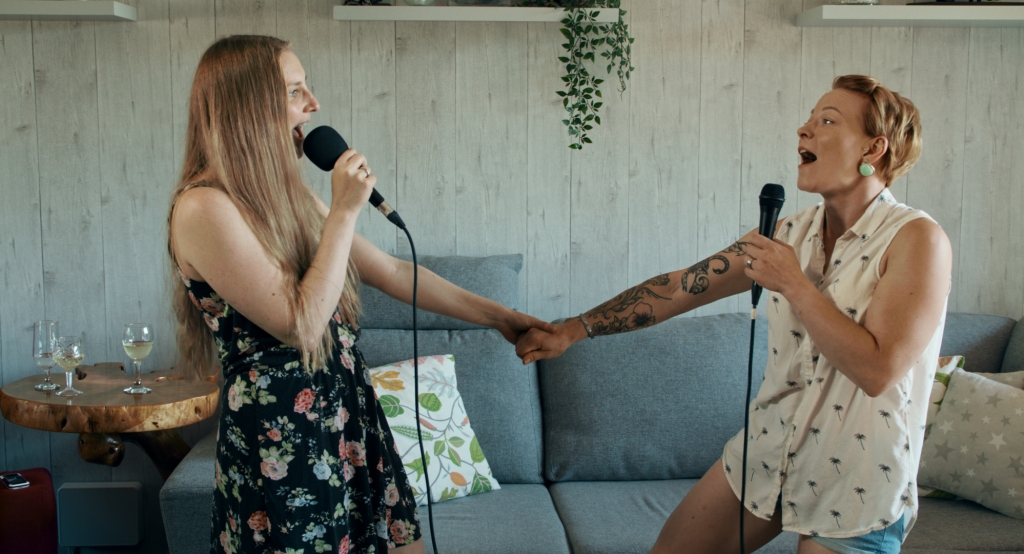 Two women standing, holding hands and singing. In the background, there is a sofa, wine glass and a house plant.
