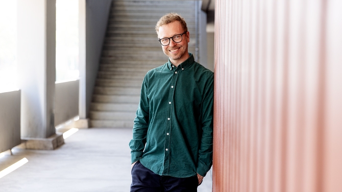 Lauri Nurkse nojaa punertavaan seinään, katsoo kameraan ja hymyilee. Taustalla näkyy ulkoportaat.