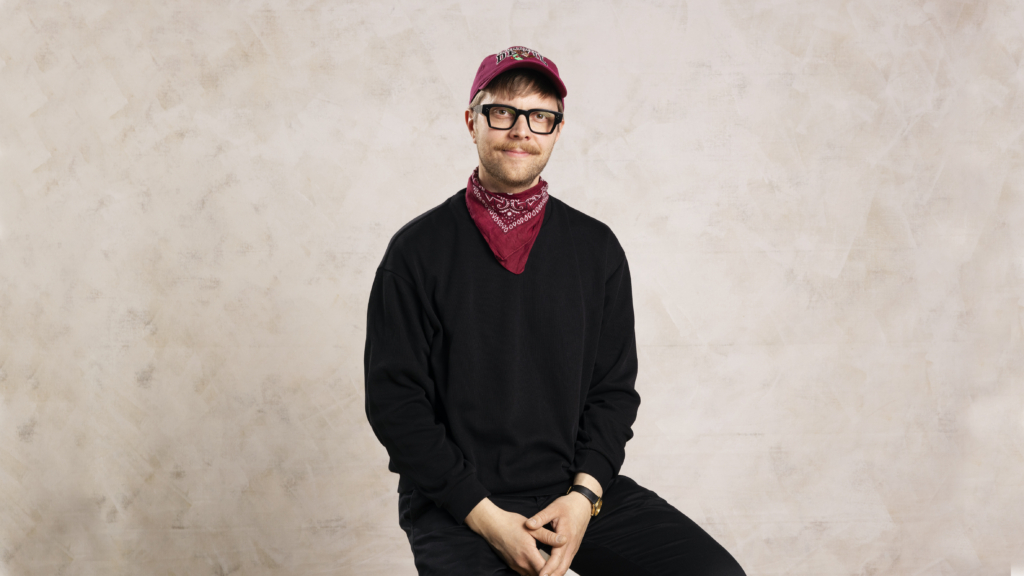 Juho-Pekka Tanskanen sits in a chair and looks at the camera. The background is light brown.