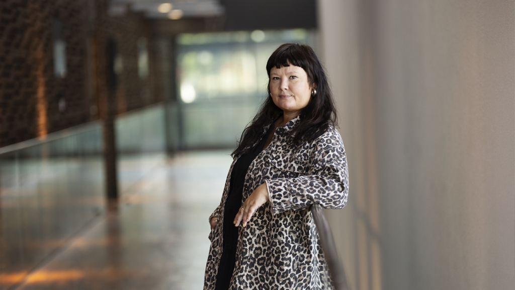 Susse Roos nojaa kaiteeseen. Takana käytävä, jonka päässä on ikkuna.